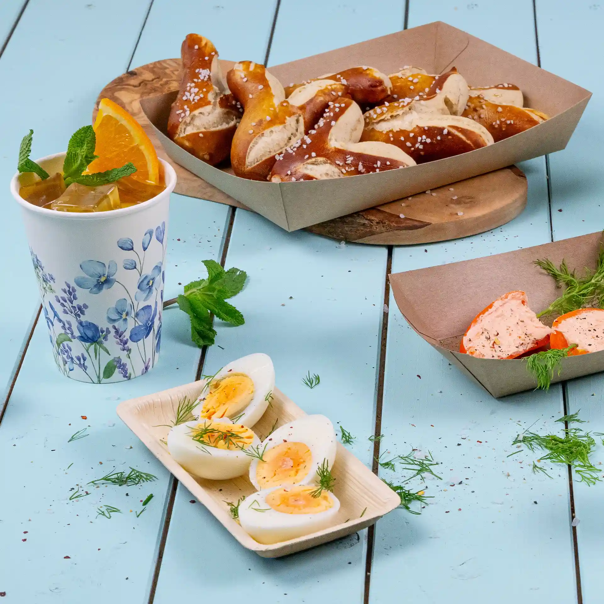 Image of a cup with a floral design, pretzels in a brown box, and sliced boiled eggs on a palm leaf tray.