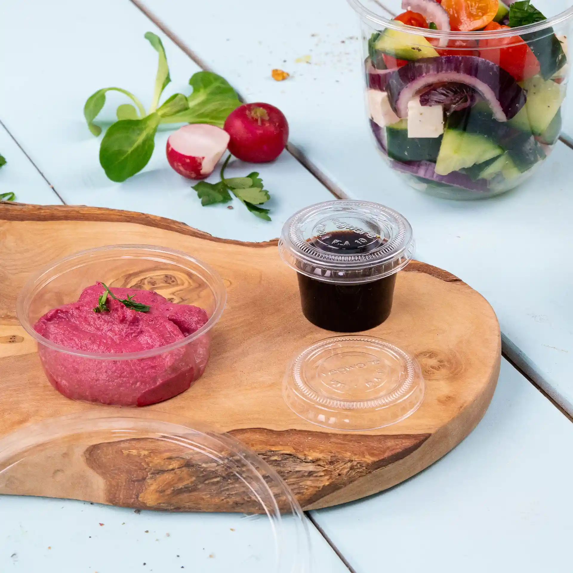 Transparent portion cups with pink dip and dark sauce on a wooden board, with salad and vegetables in the background.