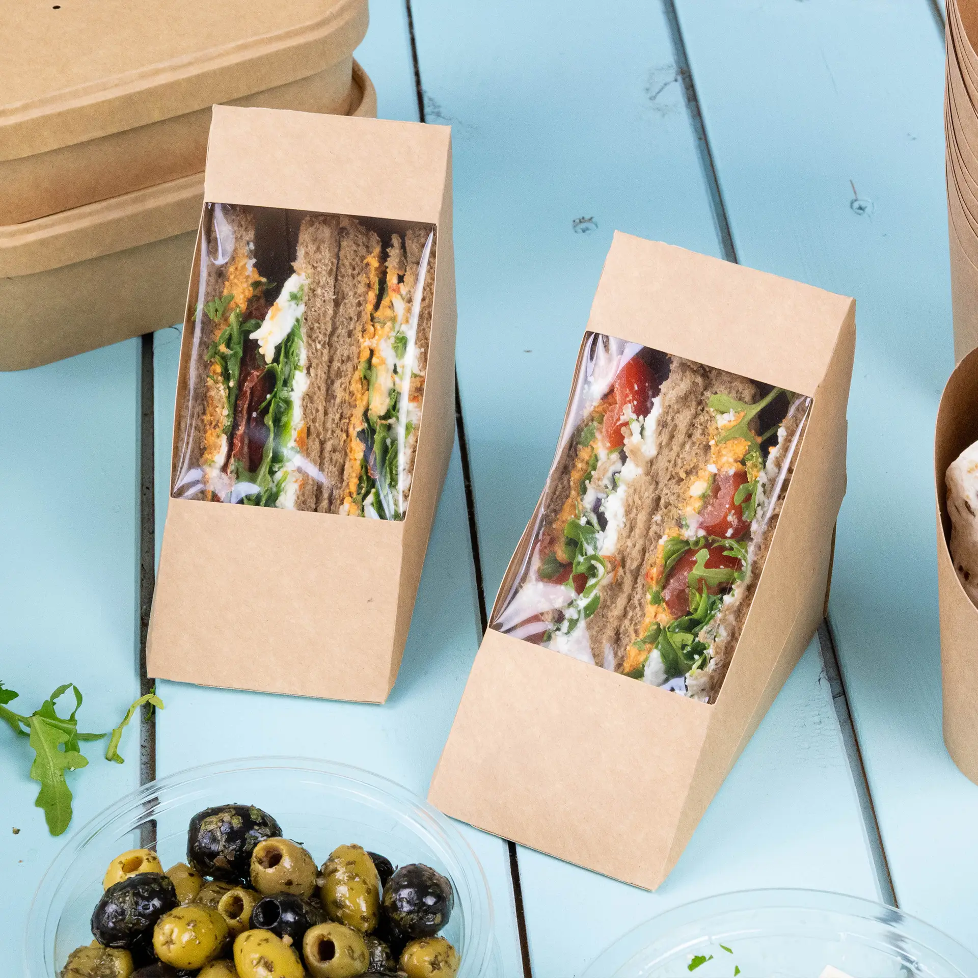 Two brown cardboard sandwich wedges with a transparent PLA window displaying sandwiches and filled rolls on a blue surface, with olives in a bowl nearby.