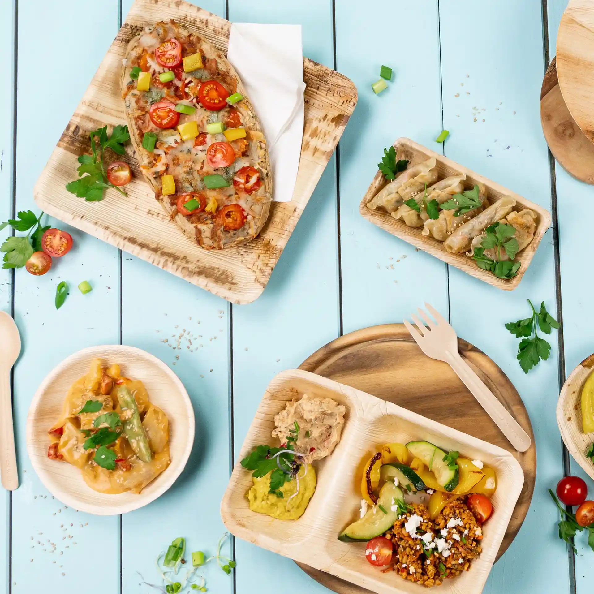 Image of various dishes served on palm leaf plates, including vegetables, sauces, and garnishes on a blue wooden table.