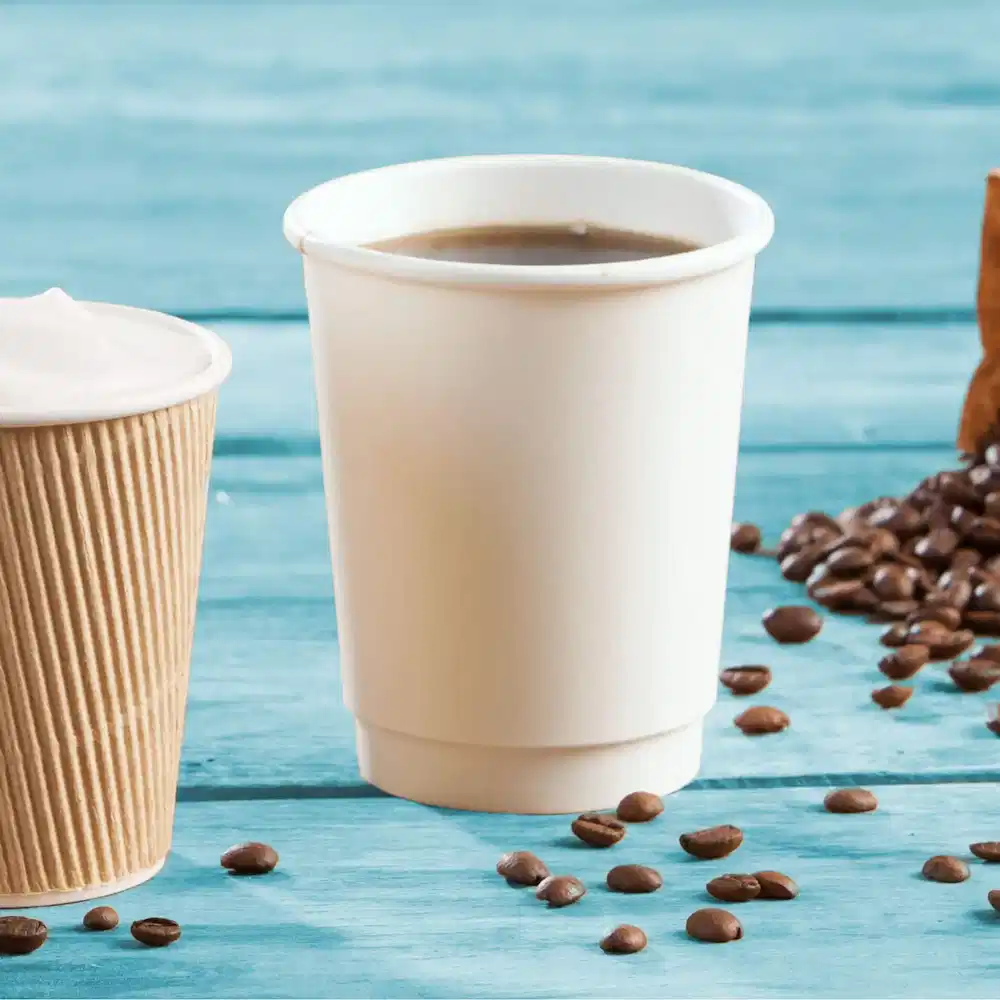 Two stacked double-walled paper cups, one plain white and one brown ribbed, with coffee beans scattered around.