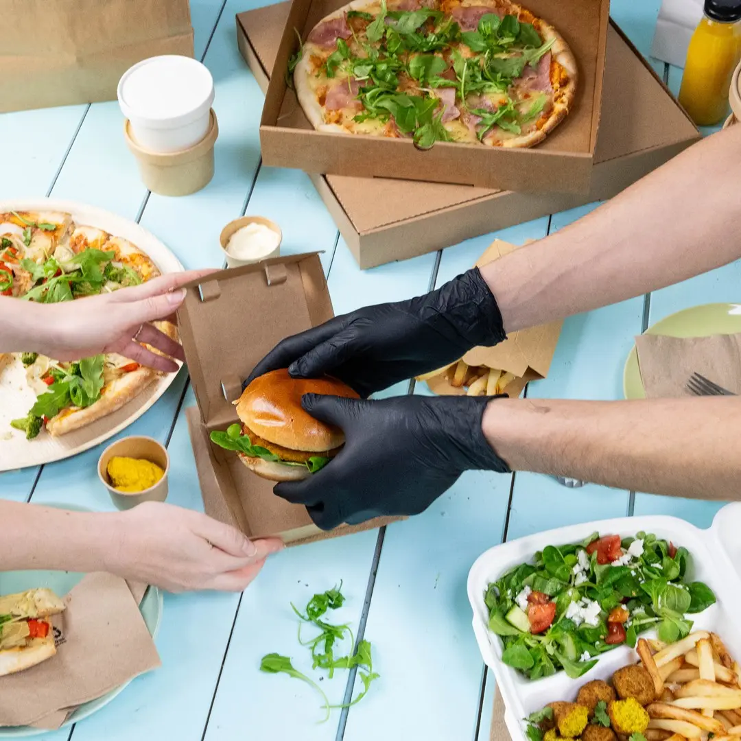 Hands in black gloves are placing a burger into a brown takeaway box, surrounded by pizza, salad, and sauces.