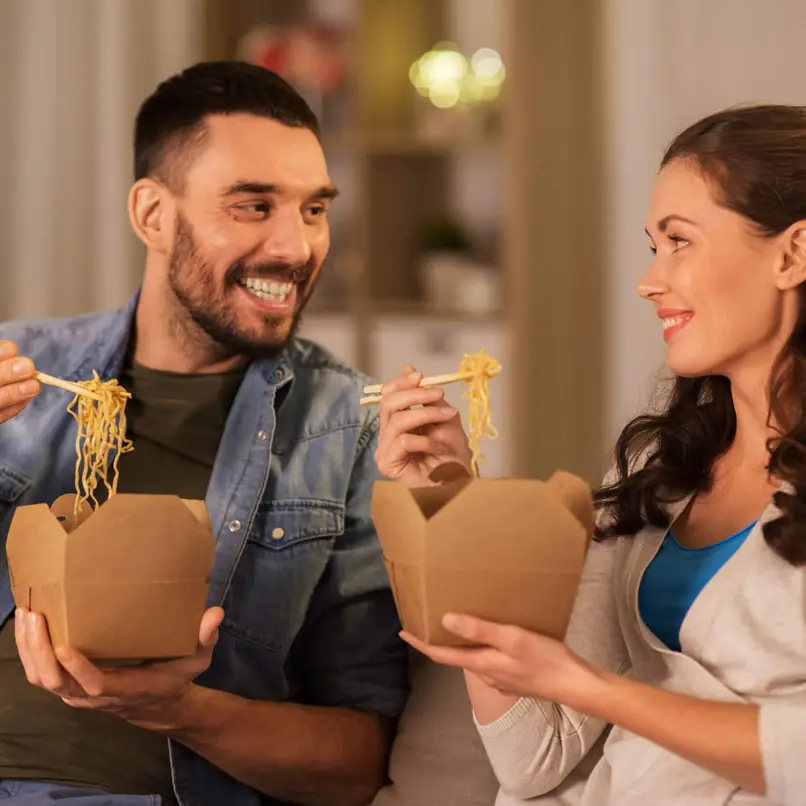 Couple enjoying takeout noodles in brown boxes, using chopsticks, sitting together in a cozy indoor setting.