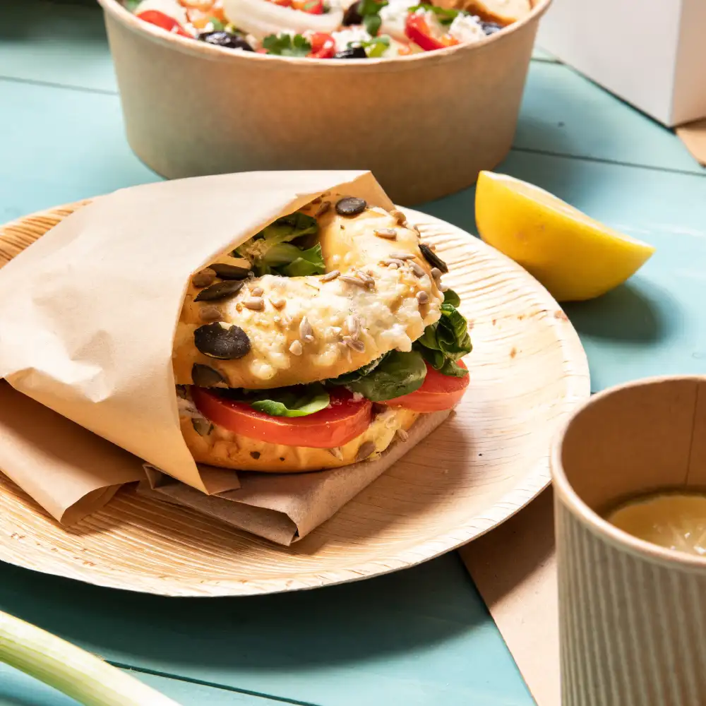 Bagel sandwich with lettuce and tomato on a wooden plate; a lemon wedge and a cup are also visible in the background.