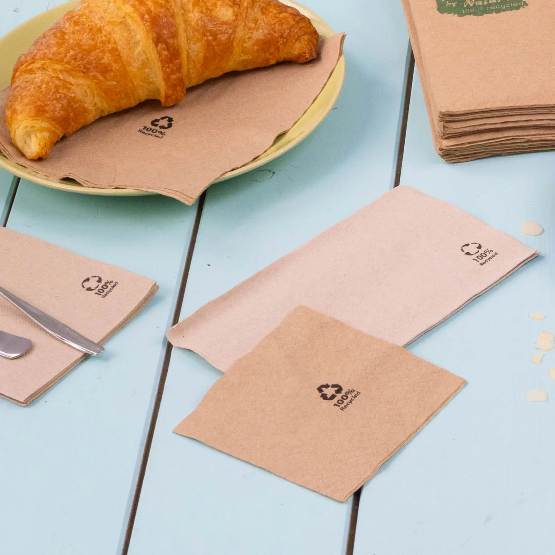 Image of brown recycled paper cocktail napkins, with subtle recycling logos, placed on a table near a croissant and cutlery.
