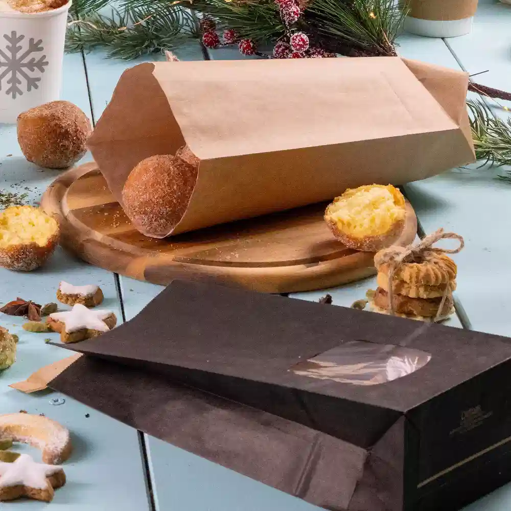 Brown paper bag with baked goods, black bag with a clear window, wooden serving board, and festive decorations.