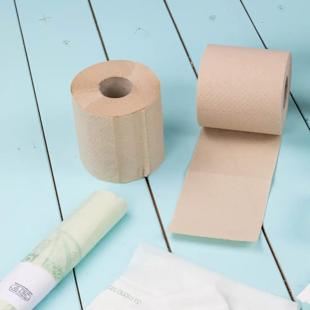 Brown paper towel roll and partially unrolled roll on a light blue wooden surface, alongside a green plastic bag and white wrapper.