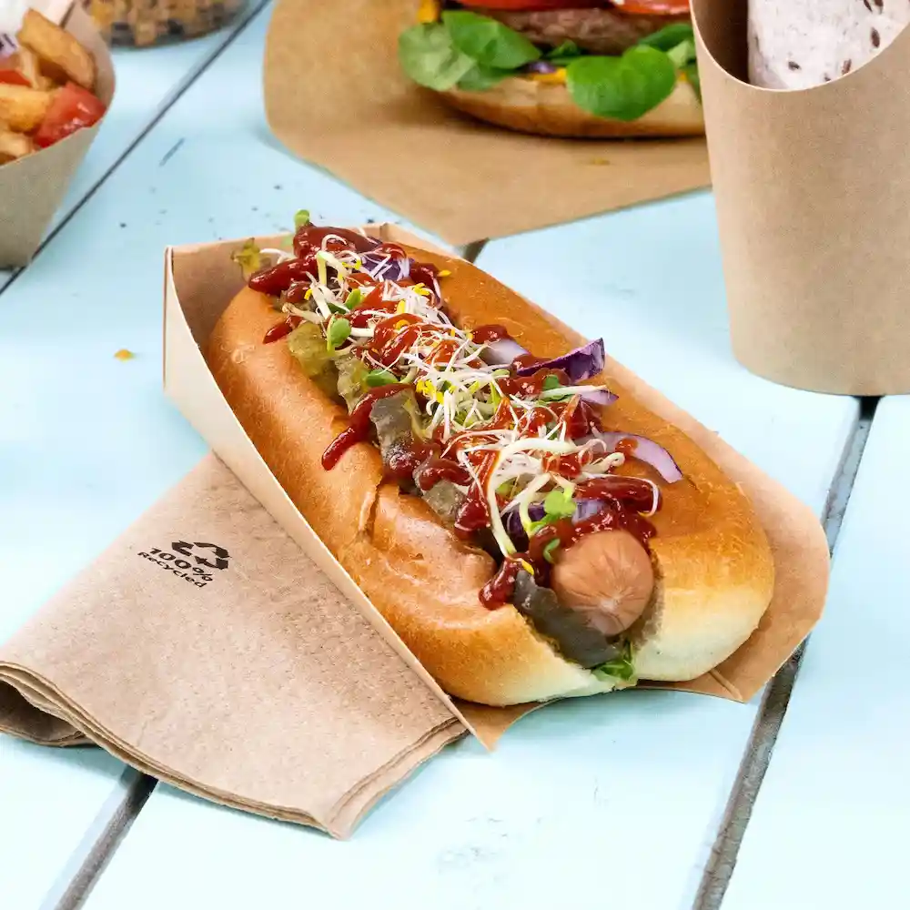 Hot dog in a brown paper holder, topped with ketchup, mustard, pickles, and sprouts, resting on a wooden table.