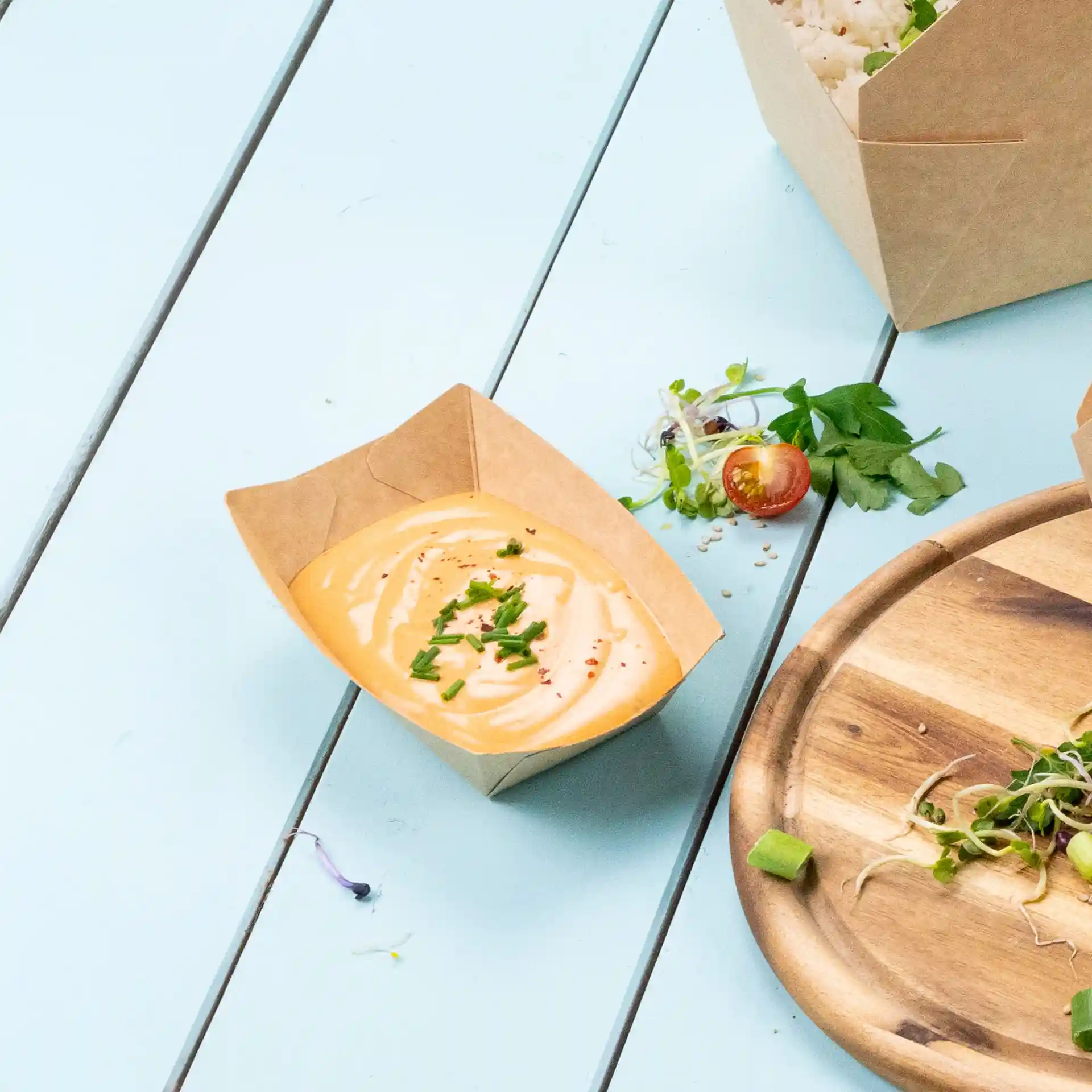 Brown cardboard snack tray with a creamy sauce topped with chopped chives, placed on a light blue surface.