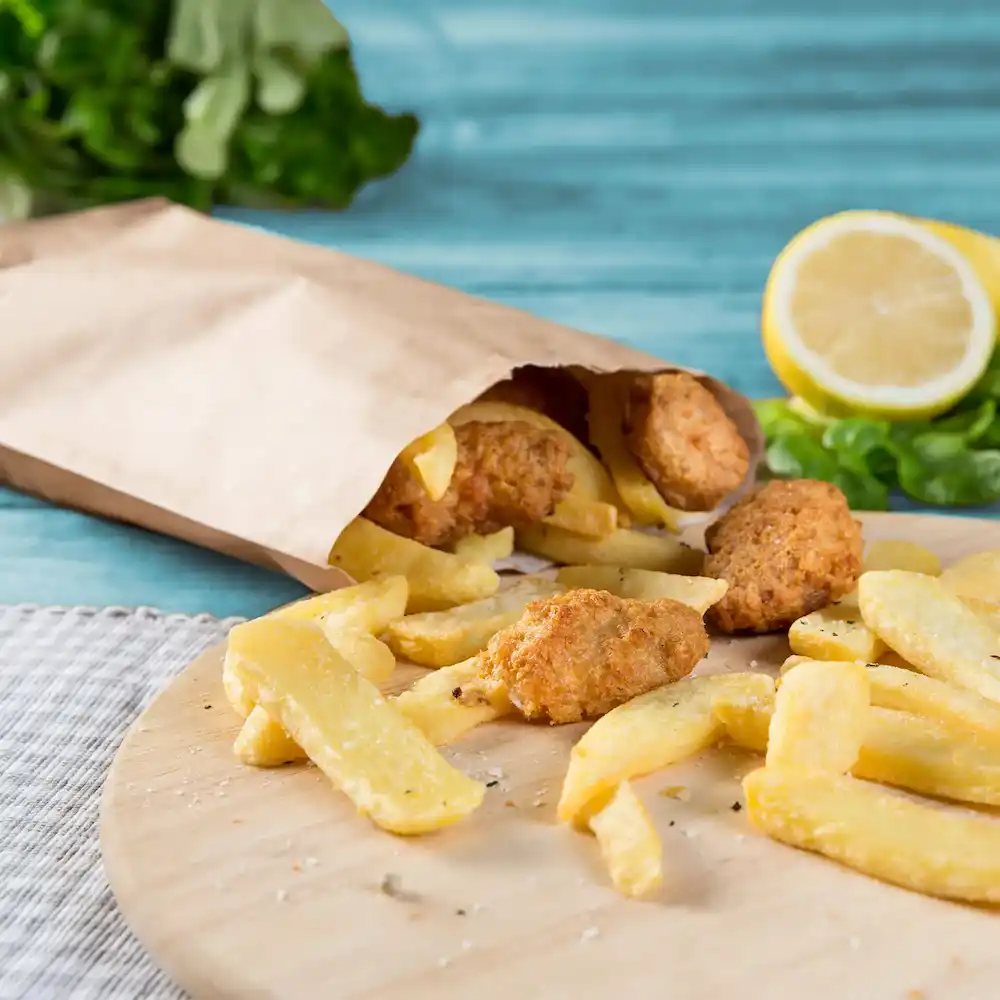 Paper bag with fried food spilling out, featuring fries and several golden-brown pieces, with lemon and greenery in the background.