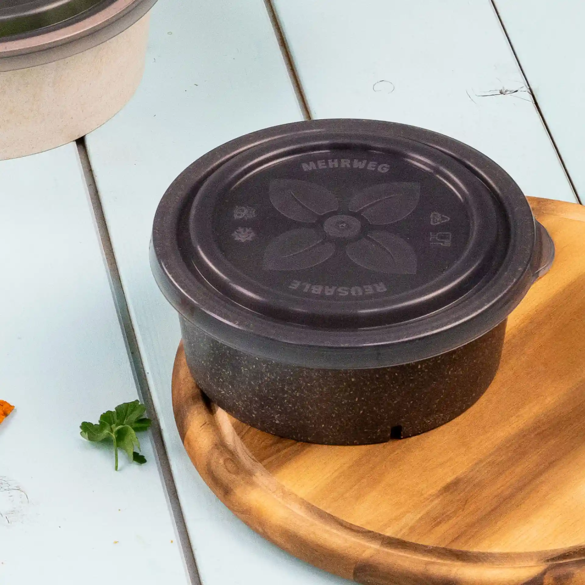 Round grey reusable bowl with a black lid, placed on a wooden surface, featuring a floral design on the lid.