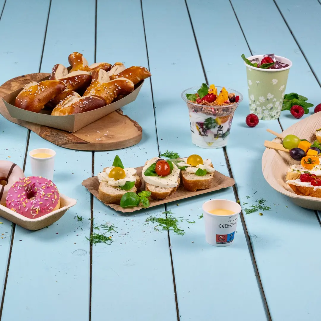 Assorted snacks including pretzels, sandwiches with toppings, yogurt parfait, donuts, and fruit skewers on a blue wooden surface.