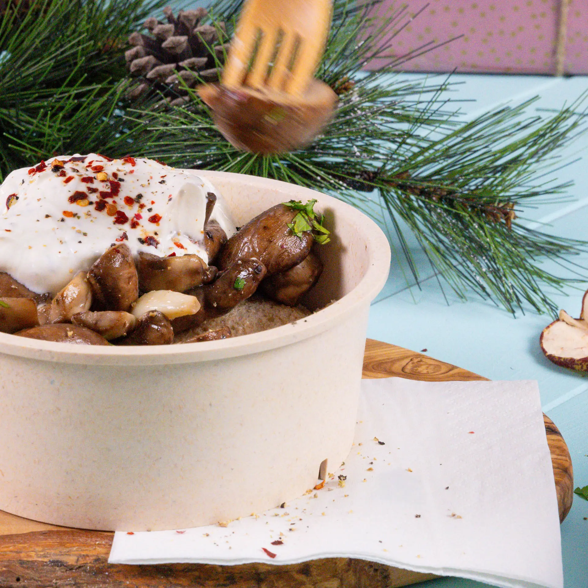 Bowl with mushrooms and yogurt topped with red pepper flakes, placed on a wooden board with a white napkin beside it.