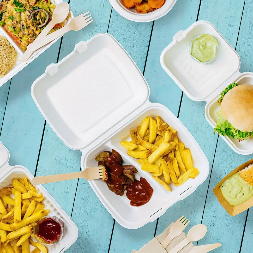 White takeaway containers with fries, sausage, and sauces on a blue wooden surface, alongside a burger and other food items.