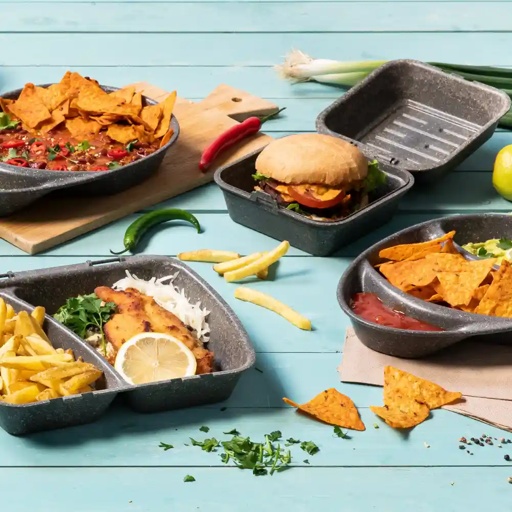 Variety of food items in reusable containers, including nachos, a burger, fries, and fish with lemon, on a blue surface.