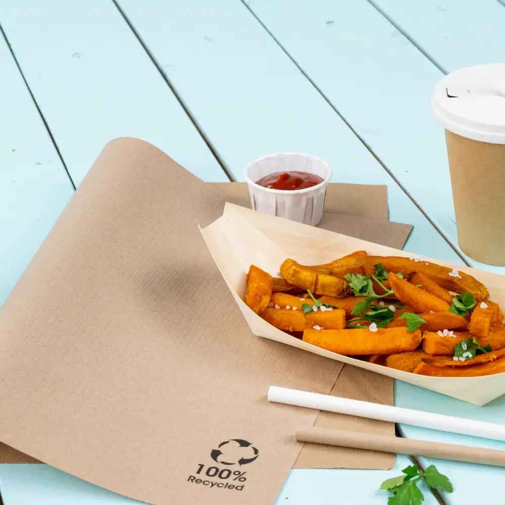 Brown kraft paper table setting with sweet potato fries in a tray, ketchup cup, a drink cup, and cutlery nearby. Recycled materials logo visible.