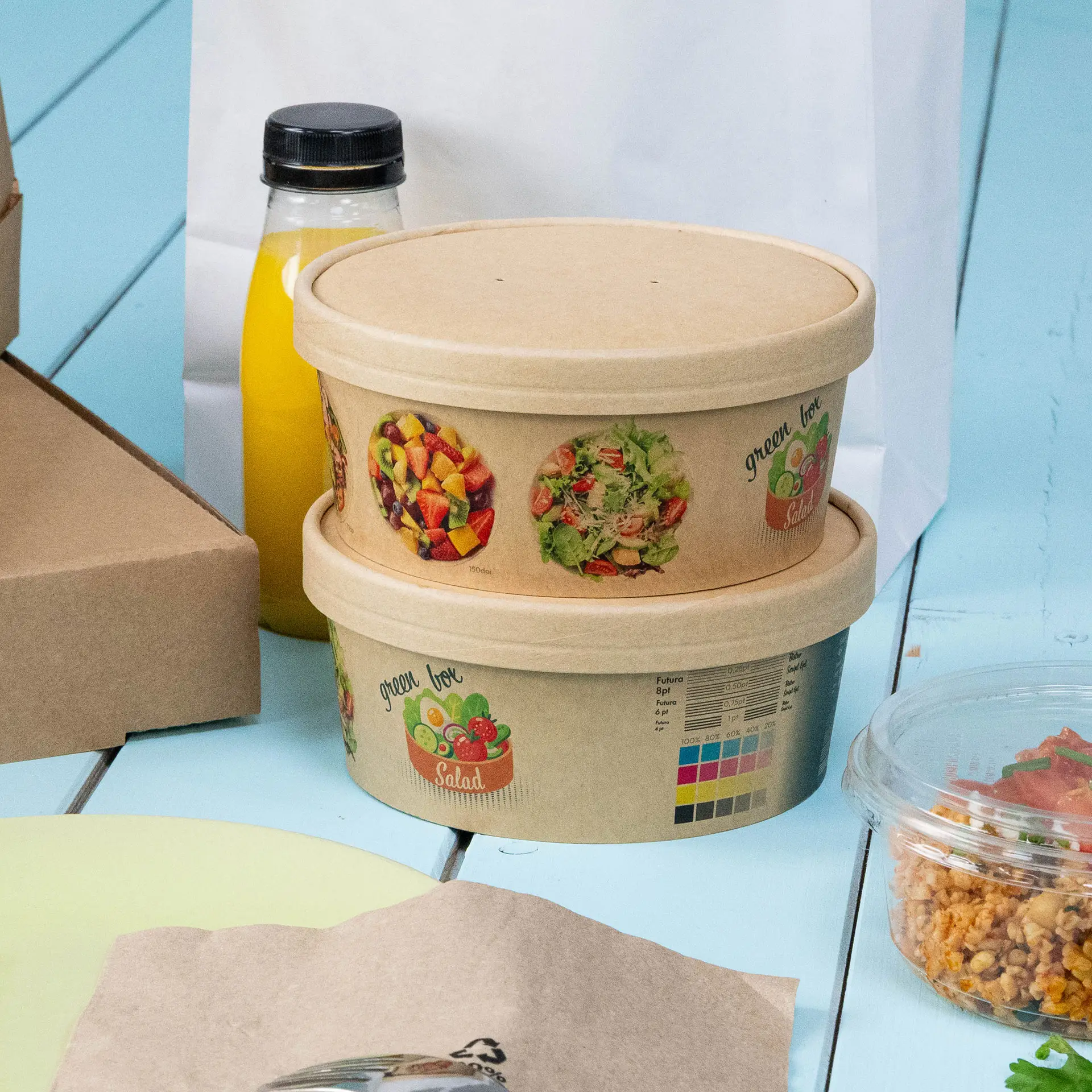 Round cardboard salad bowls with custom prints; shown stacked; includes a yellow drink and additional food containers nearby.