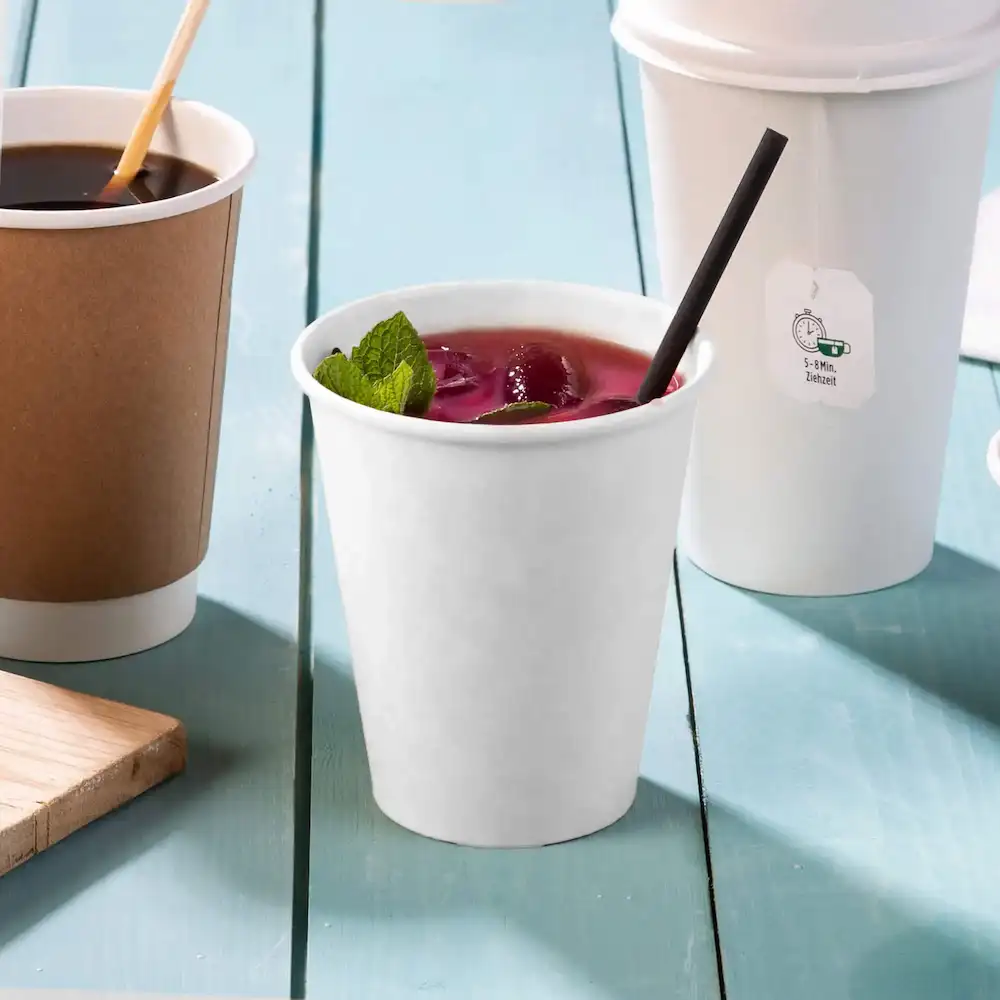 White paper cup containing a beverage with mint leaves, black straw, and two other cups in the background.