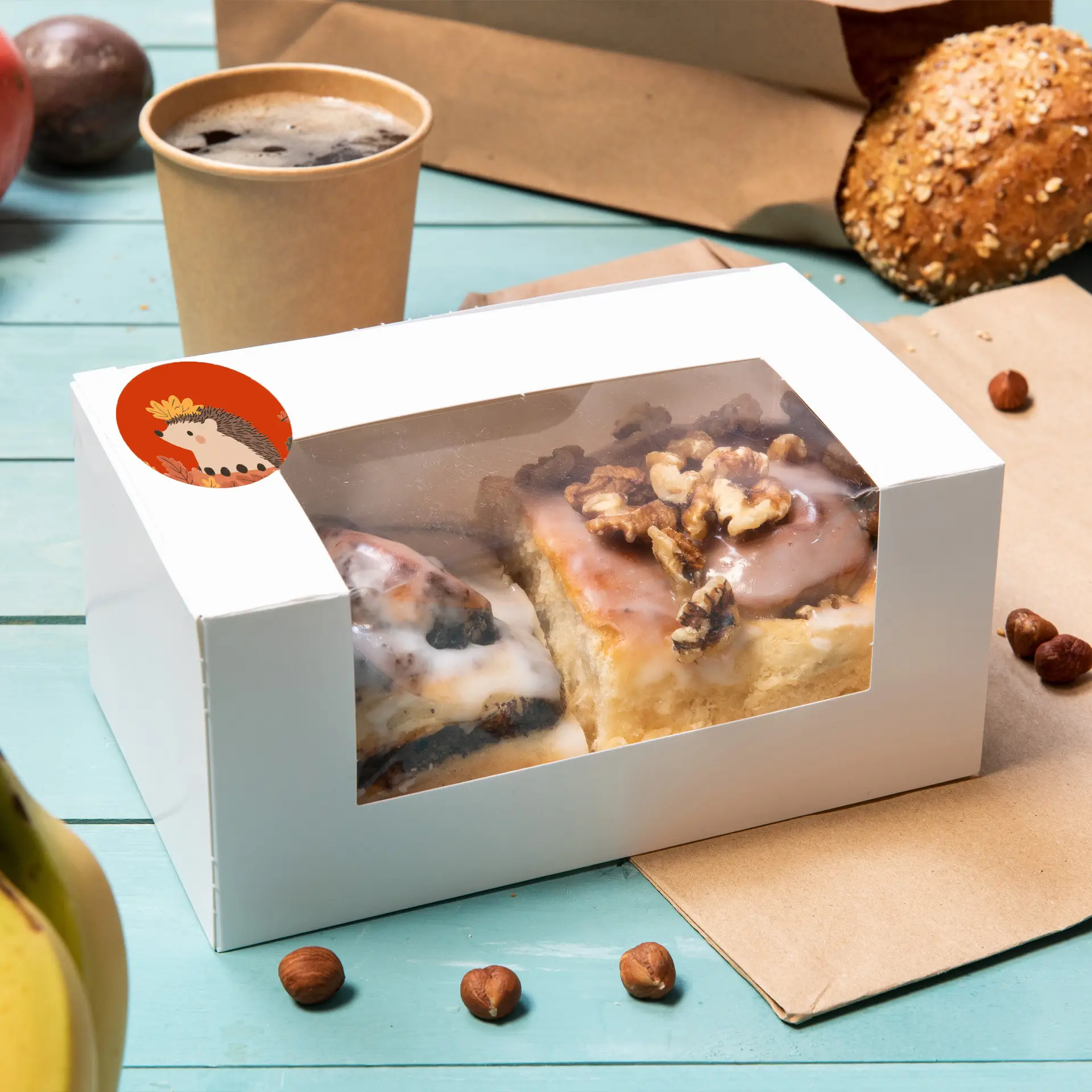 Two pastries in a clear window box, one with icing and the other topped with walnuts, next to a coffee cup and scattered nuts.