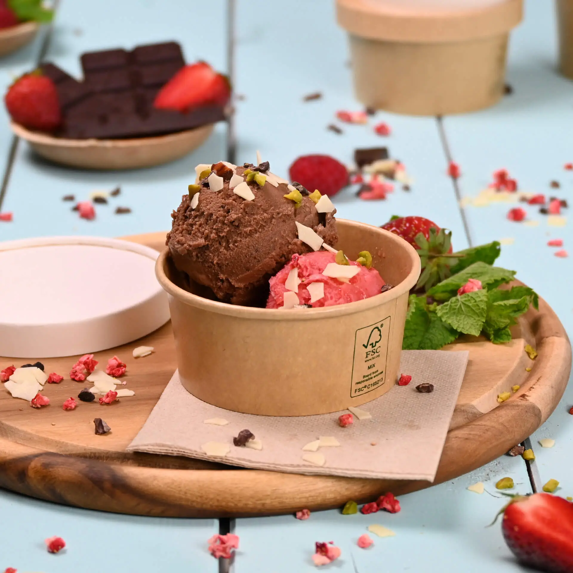 Brown paper ice cream cup containing chocolate and strawberry ice cream, topped with nuts and surrounded by fresh strawberries and mint.