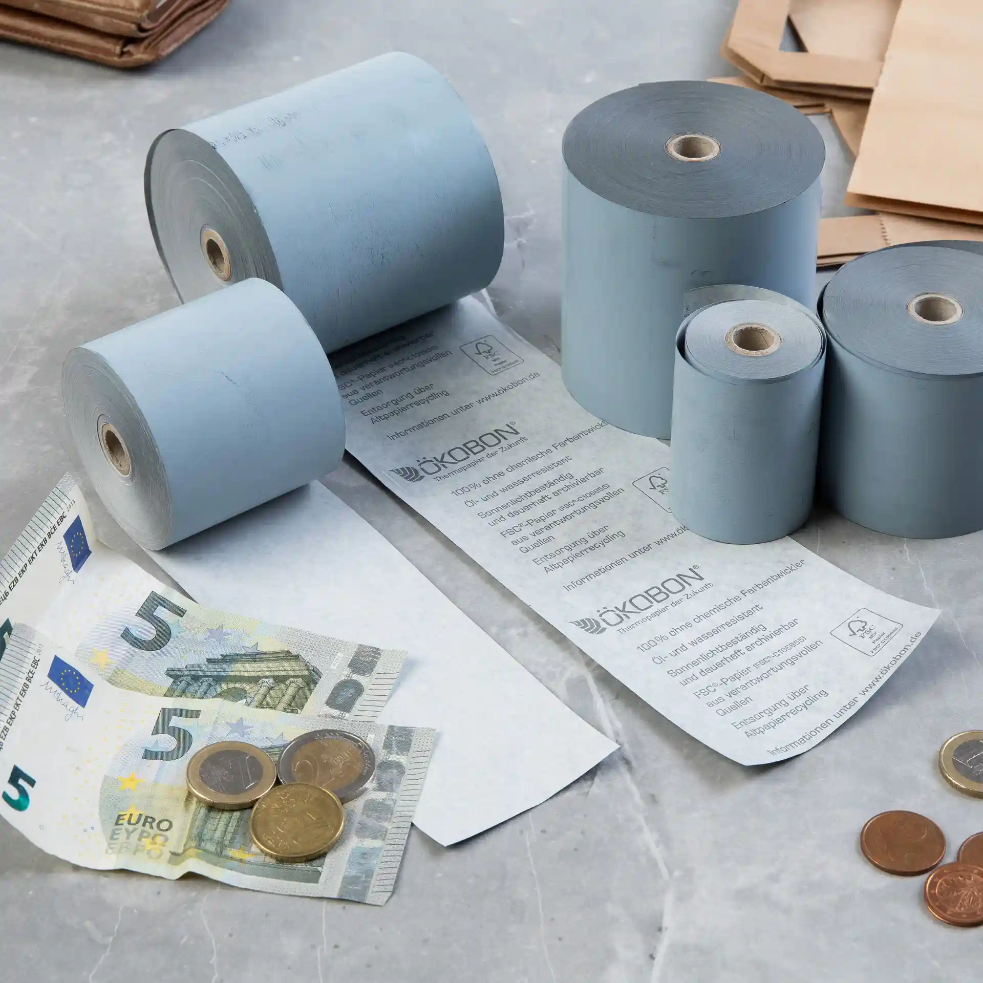 Image of several grey thermal paper rolls, a 5 euro banknote, and coins on a surface.