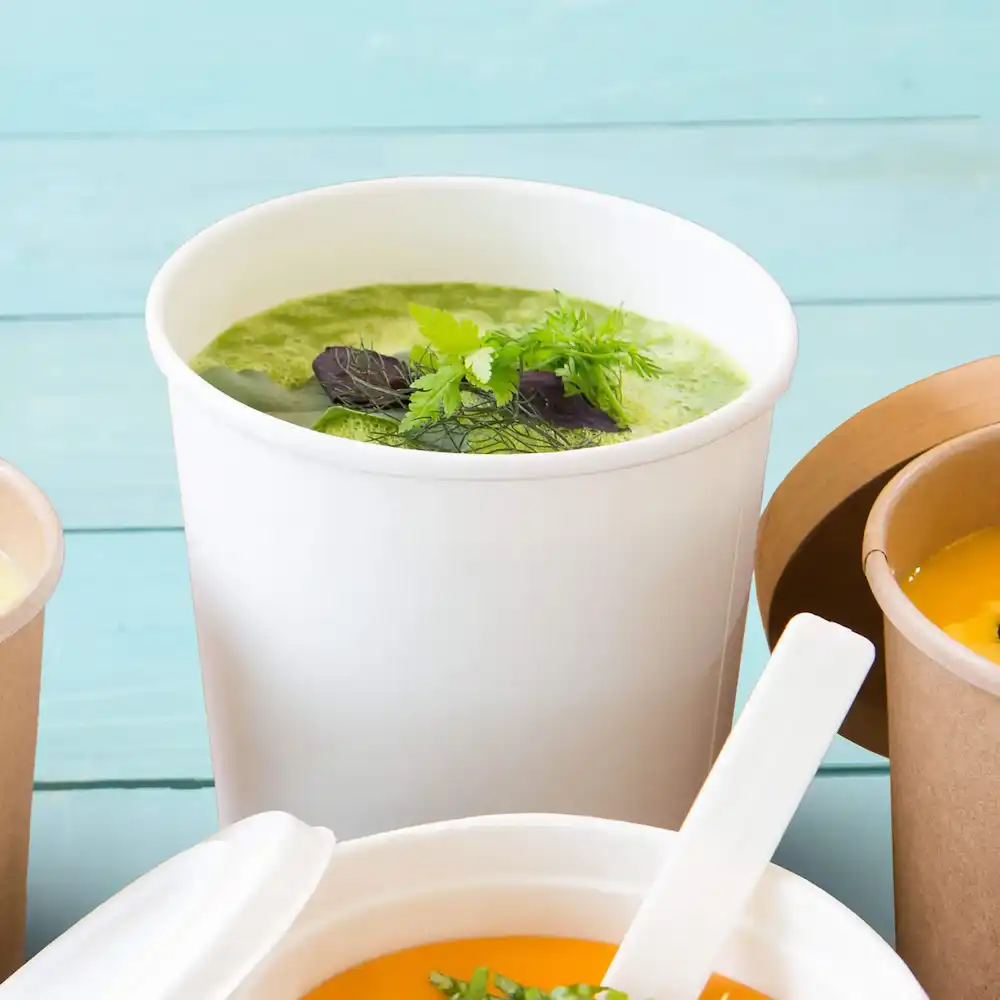 White soup cup filled with green soup and garnished with herbs, surrounded by other containers.