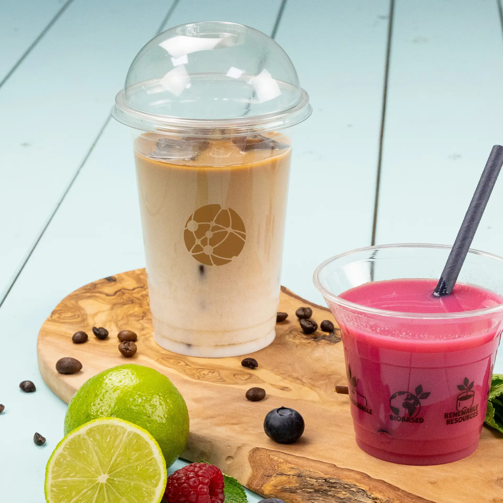 One clear PLA cup filled with iced coffee, labeled with a logo; next to it, a smaller cup with pink drink and a straw, on a wooden board.