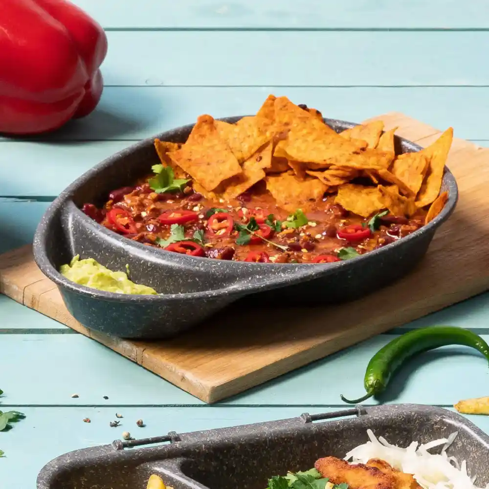 Bowl with chili topped with tortilla chips, sliced red chilies, and cilantro on a wooden board with toppings.