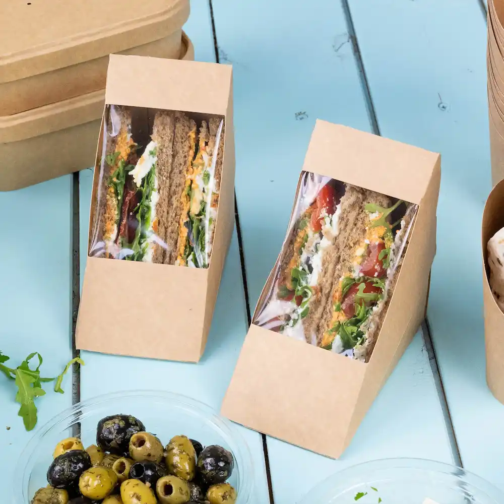 Two sandwich boxes made of brown kraft paper with window displays of layered sandwiches and a bowl of mixed olives.