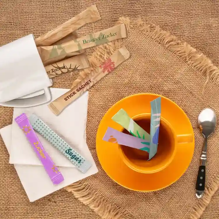 Assorted sugar stick sachets placed in a cup of tea on a burlap surface, with a spoon and napkin nearby.