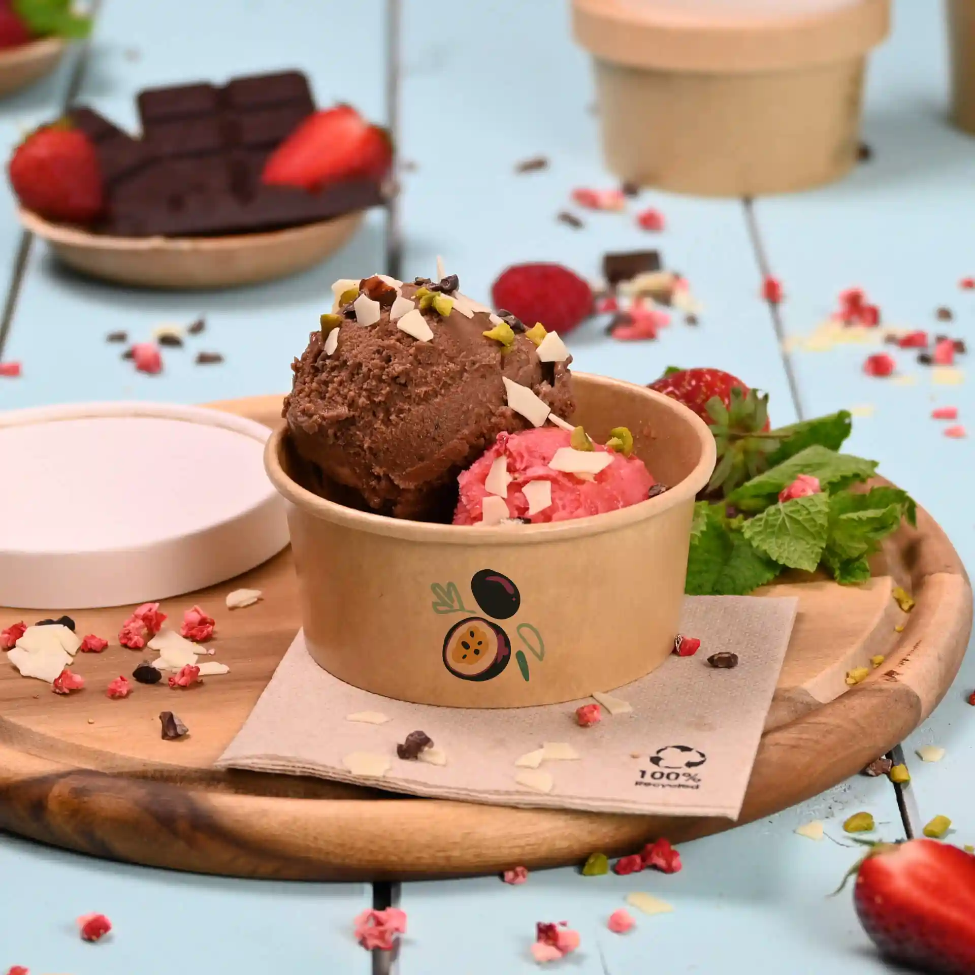 Round brown ice cream cup with two scoops of ice cream, topped with nuts, on a wooden board with strawberries and mint.