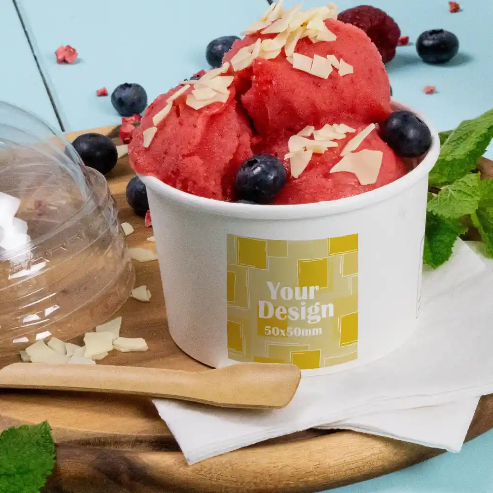 Cup of red sorbet topped with almonds and blueberries, labeled "Your Design 50x50mm," on a wooden surface with a spoon.