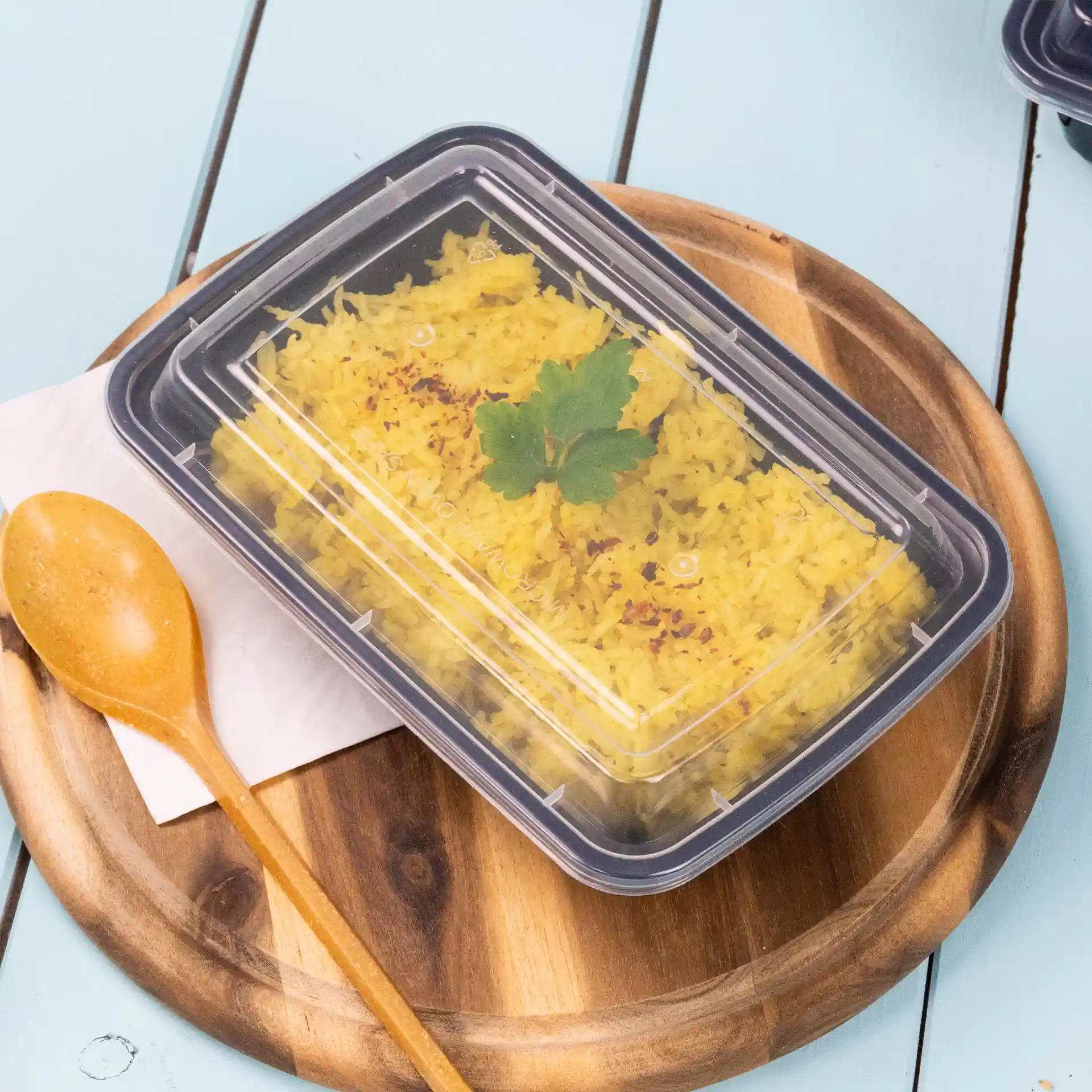 Transparent rectangular container with a reusable lid, holding yellow rice and garnished with a cilantro leaf, on a wooden plate.