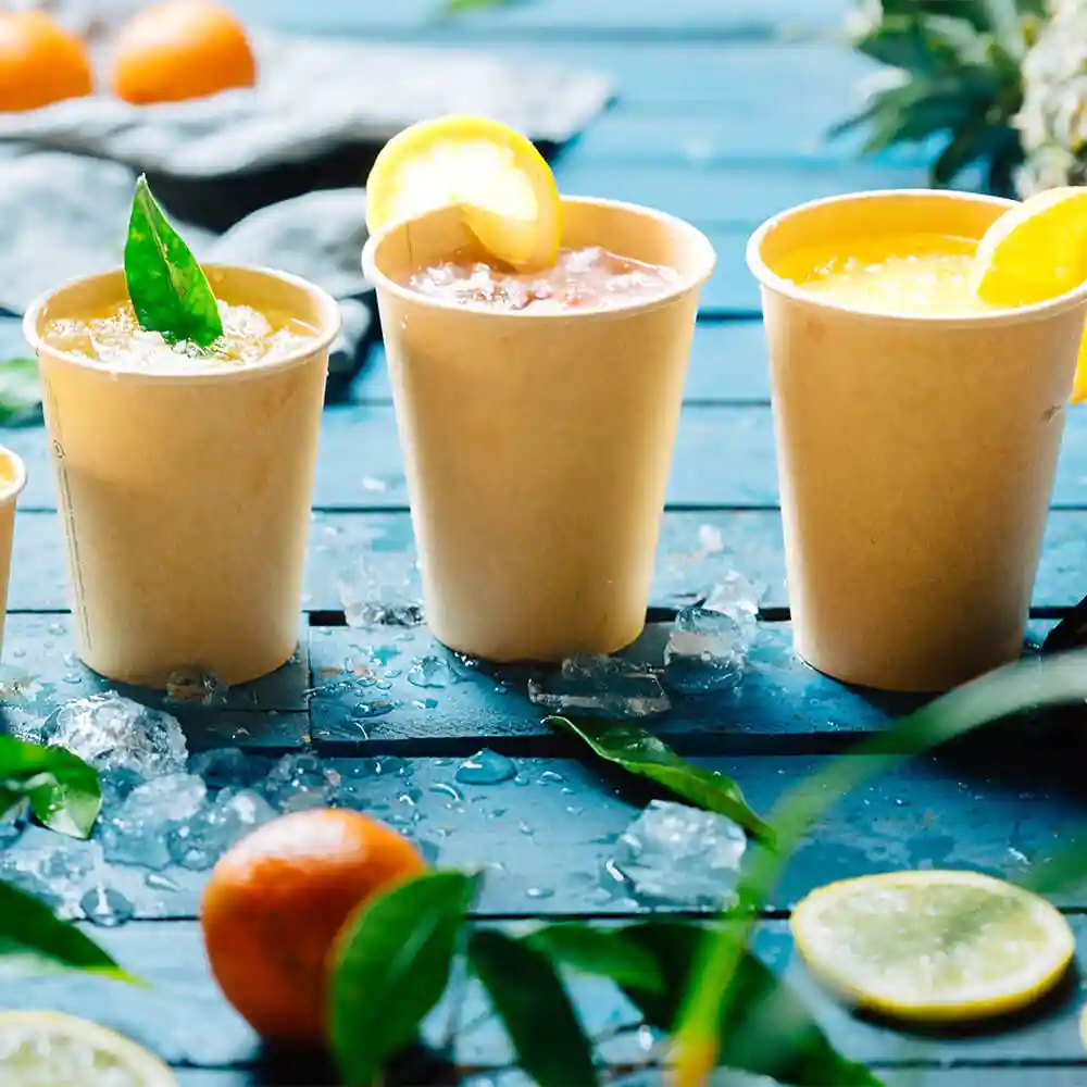 Three brown 12 oz paper cups filled with drinks, garnished with lemon and leaves, on a blue wooden surface with ice.
