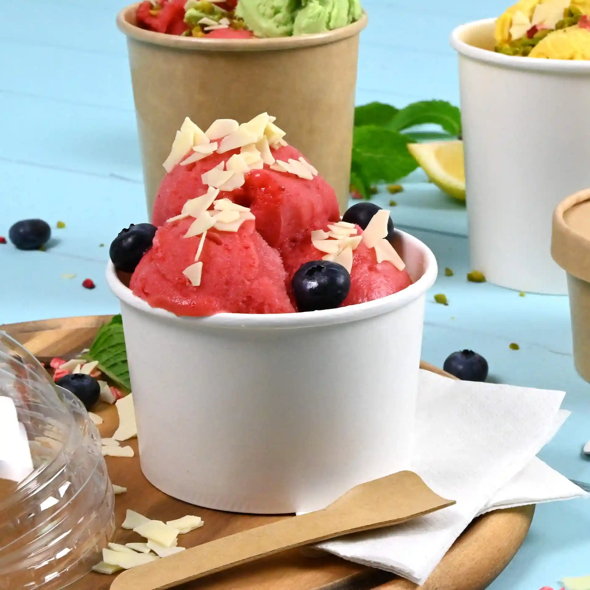 White paper cup filled with pink ice cream topped with almond flakes and blueberries, placed on a wooden surface.