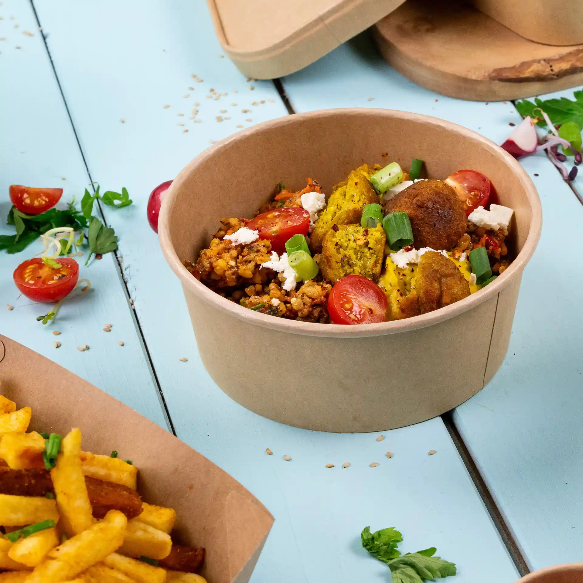 Round brown paper bowl filled with rice, vegetables, and cherry tomatoes, placed on a blue surface with fries nearby.