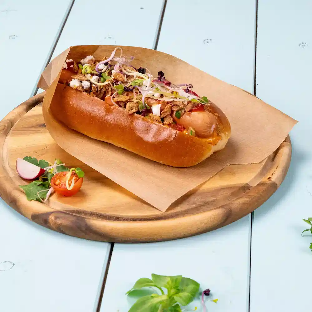 Hot dog in a bun topped with onions, herbs, and crunchy toppings on a wooden platter with side vegetables.