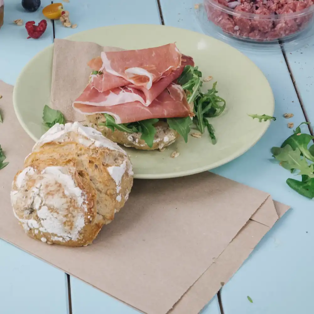 Alt text: Sliced prosciutto atop a green plate with arugula on a brown bun; a round, flaky bread roll is nearby.
