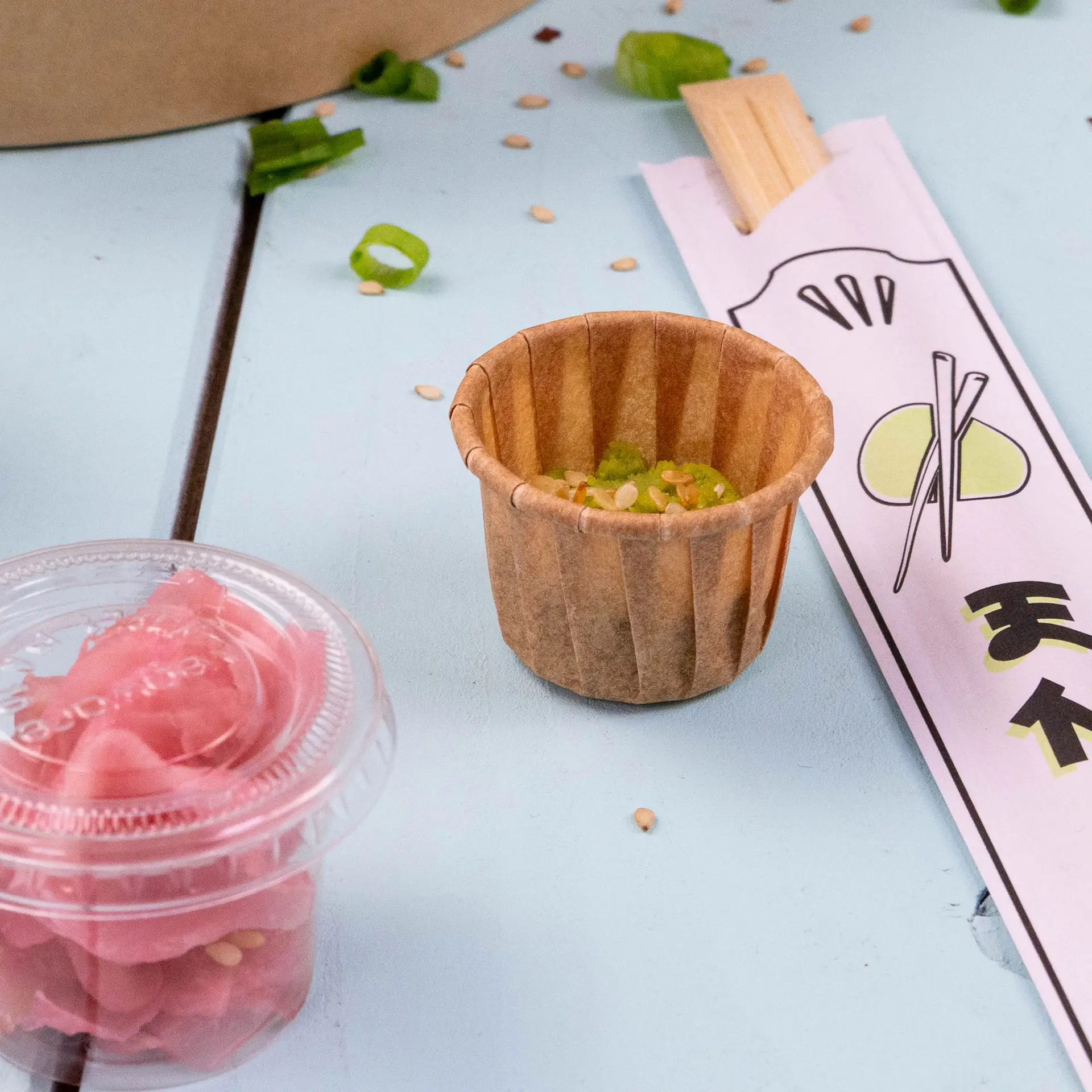 Brown paper dip cup with green garnish and sesame seeds, accompanied by pickled ginger and a chopstick wrapper on a blue surface.