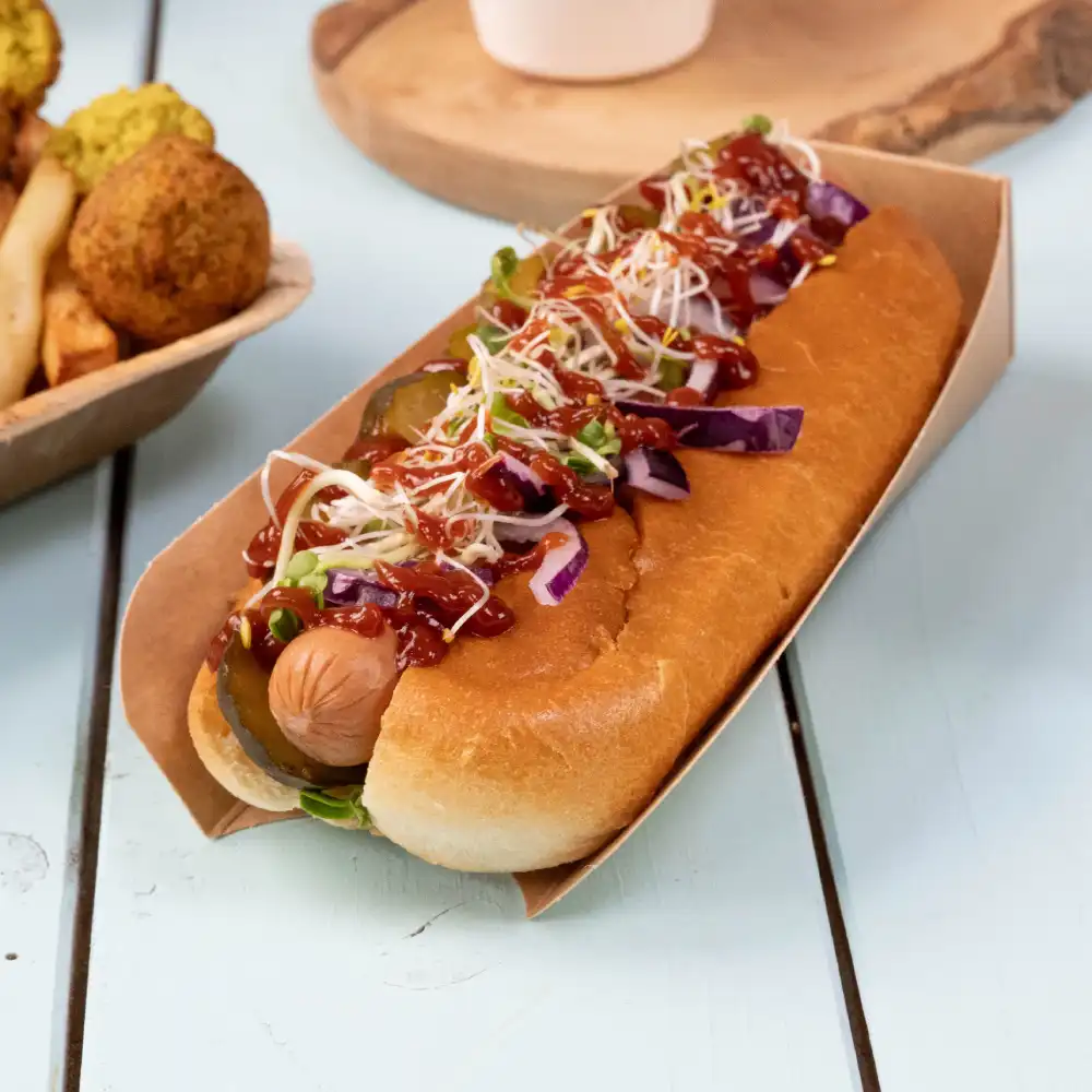 Hot dog in a paper holder, topped with pickles, shredded cabbage, onions, and ketchup, served with side items.