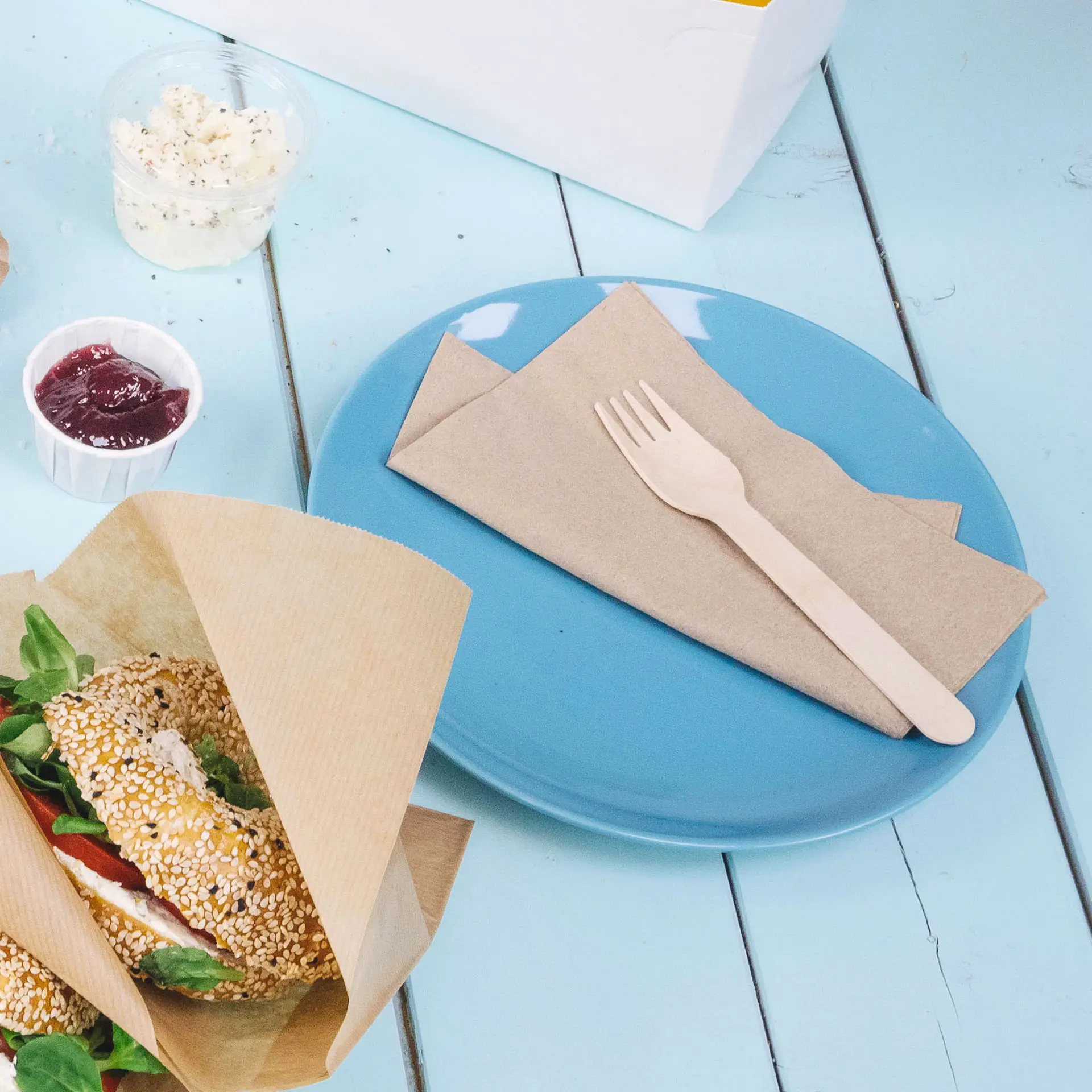 Blue plate with two unbleached paper napkins, a wooden fork, a sandwich wrapped in paper, and a small cup of jam.