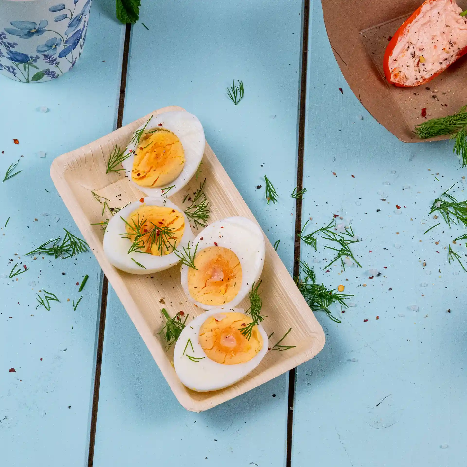 Rectangular palm leaf bowl with four halved boiled eggs garnished with dill, placed on a blue wooden surface.