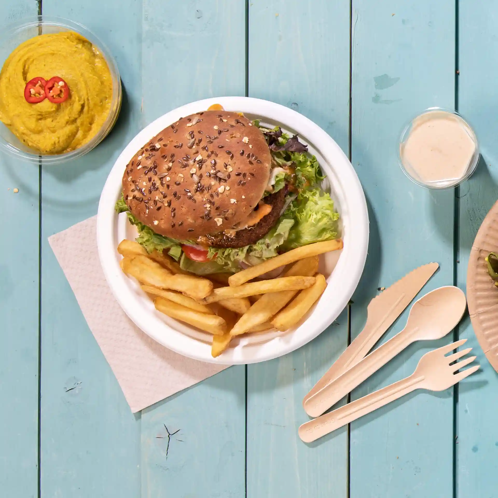 Image of a round white sugarcane plate with a burger, fries, and a bowl of hummus on a blue wooden surface.