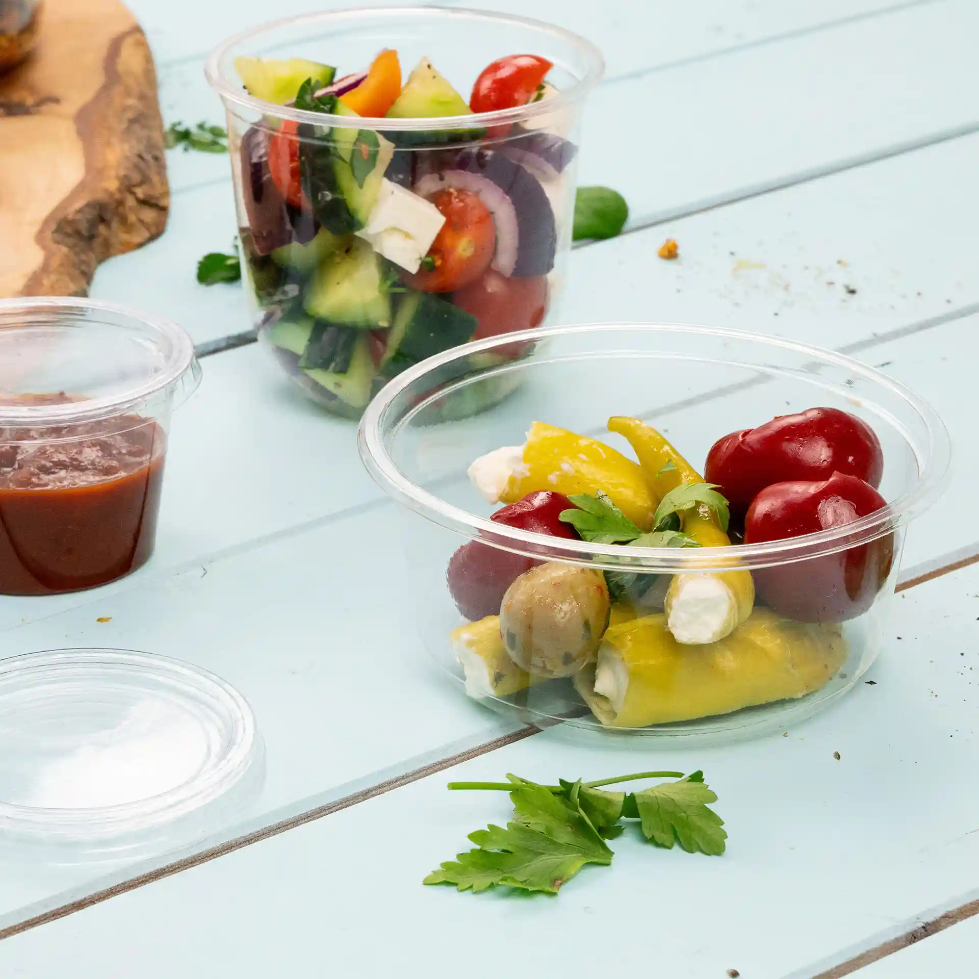 Transparent round deli container with assorted vegetables, olives, and herbs on a blue wooden surface.