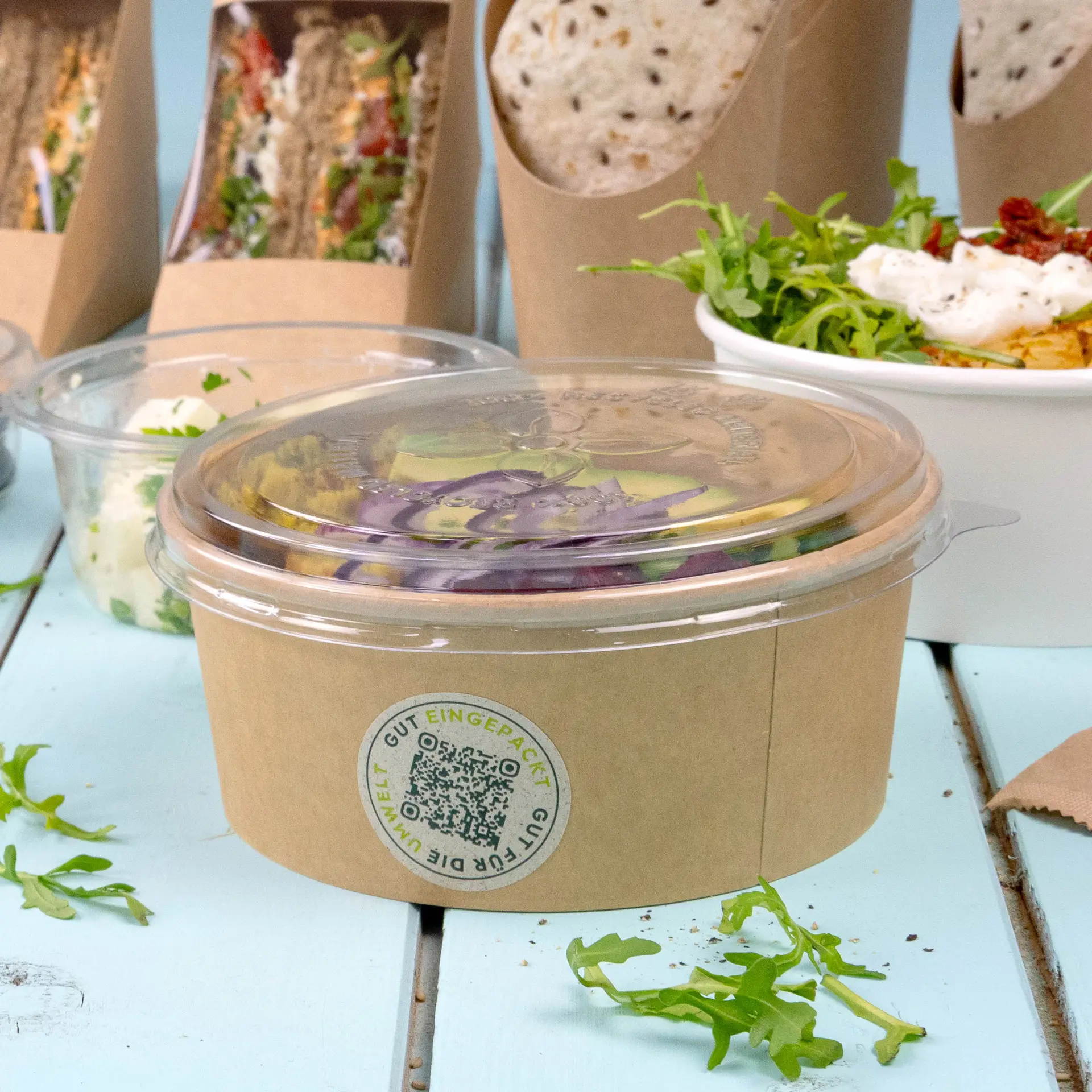 Clear rPET lid for a PLA deli container; coffee-colored base; surrounded by various food items and a small amount of arugula.