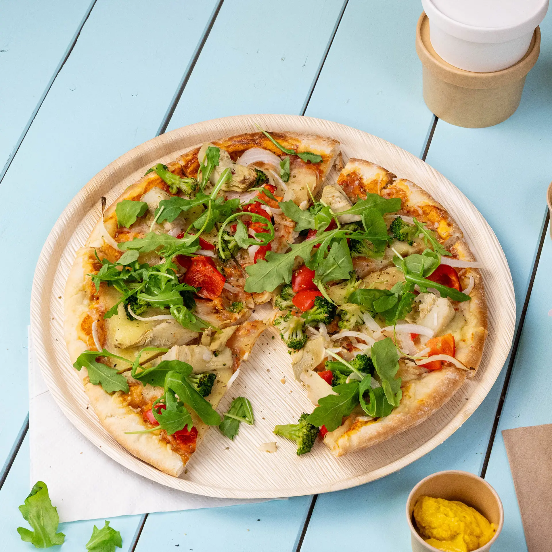 A pizza on a palm leaf plate, topped with vegetables and arugula, next to a small container of mustard.