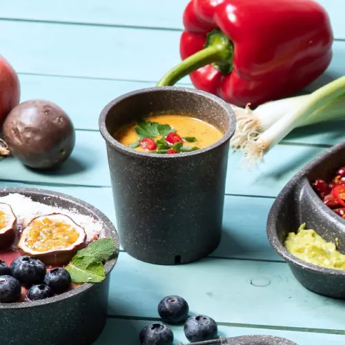 Reusable soup cup filled with soup and garnished with herbs, surrounded by fresh vegetables and fruit on a blue surface.