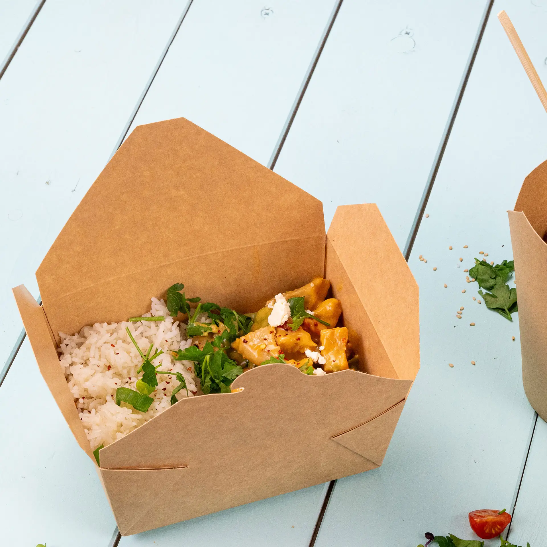 Brown cardboard take-out box filled with rice and stir-fried food, on a light blue surface, surrounded by fresh herbs.