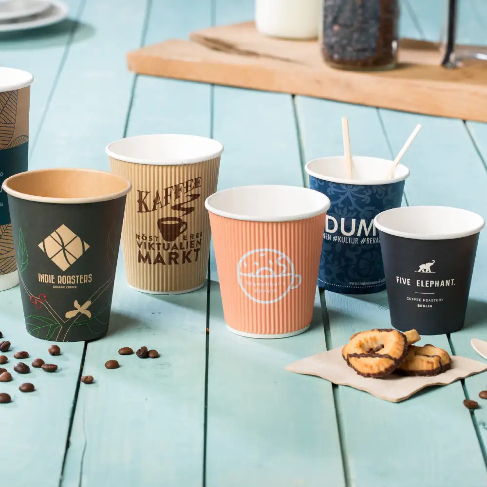 Several textured paper cups with various designs are arranged on a wooden surface, accompanied by cookies and coffee beans.