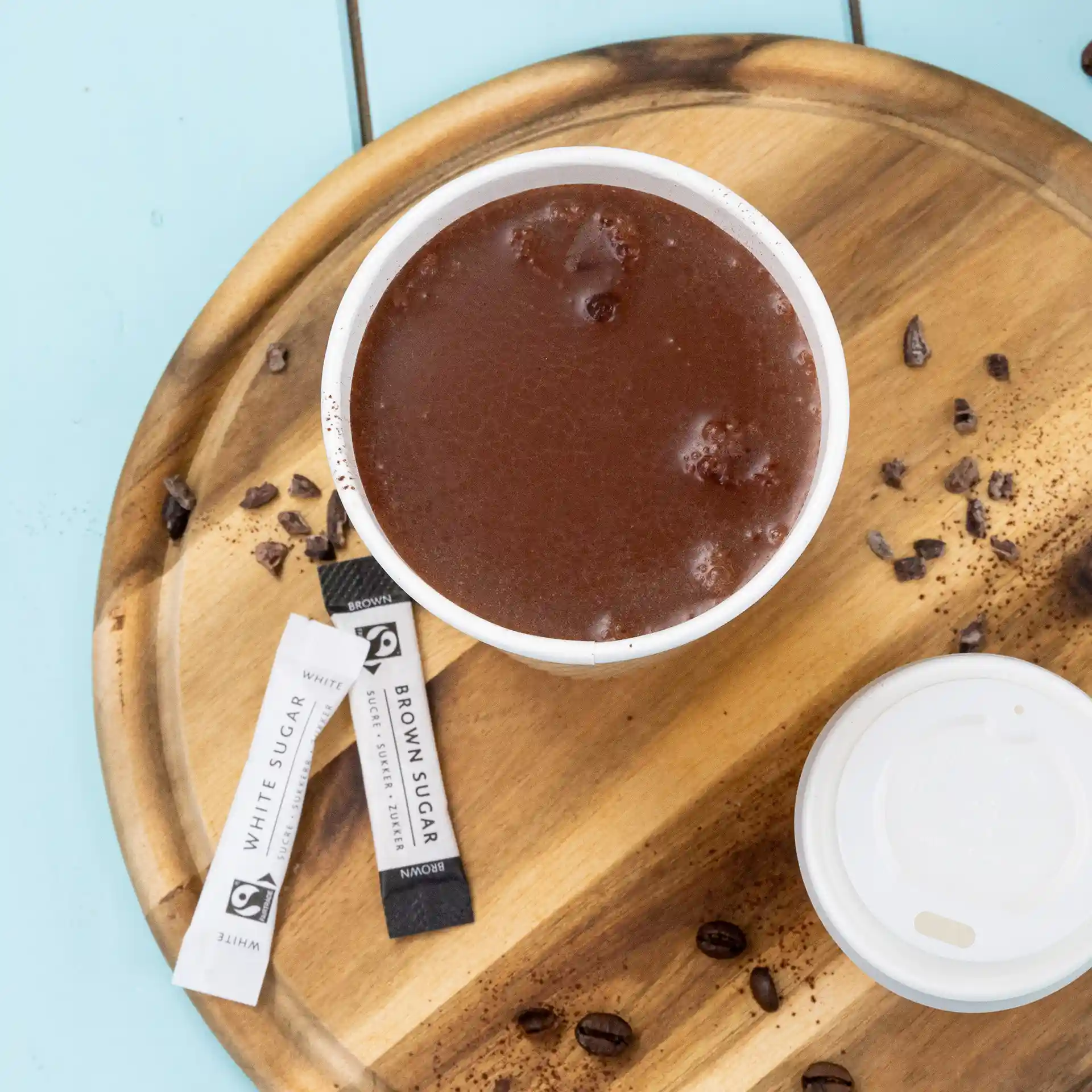 Cup of hot chocolate on a wooden tray with sugar packets and coffee beans scattered around.